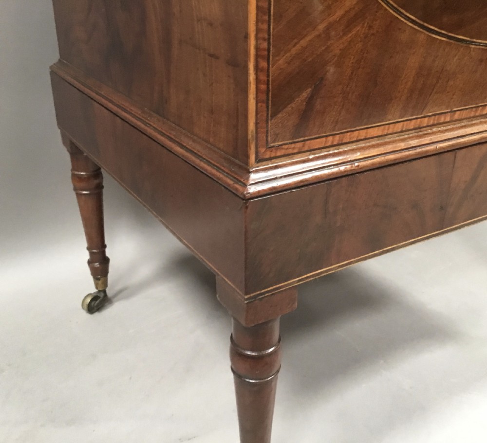 A George III Sheraton style flame mahogany cellarette featuring a domed lid, oval stringing inlay, brass side handles, and turned legs on castors. View V