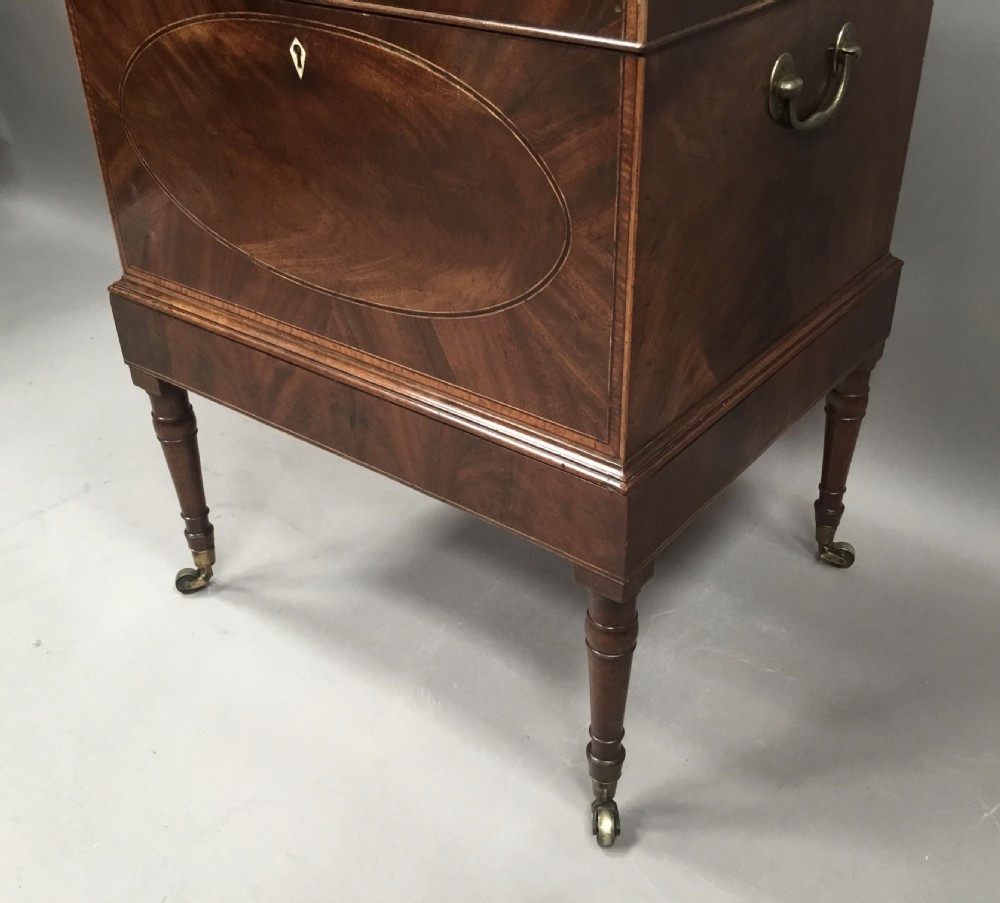 A George III Sheraton style flame mahogany cellarette featuring a domed lid, oval stringing inlay, brass side handles, and turned legs on castors. View XVI