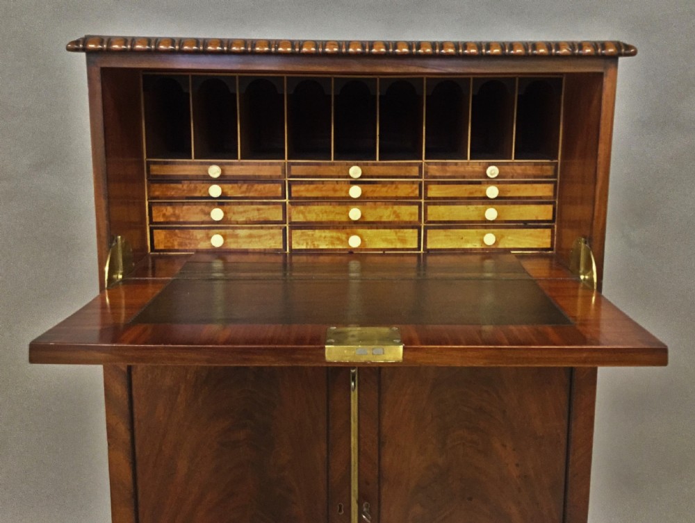A Regency period mahogany secretaire cabinet featuring flame mahogany veneered doors with re-entrant corners, gadrooned borders, and bracket feet. View V