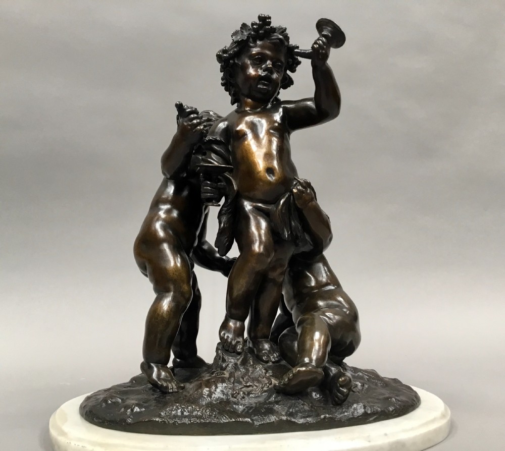 A nineteenth-century dark bronze sculpture depicting three Bacchanalian putti with grape clusters and cups, mounted on a circular white marble plinth. View VII