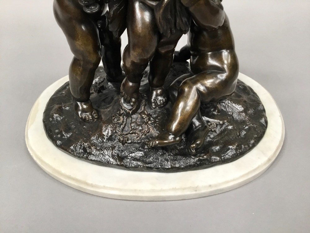 A nineteenth-century dark bronze sculpture depicting three Bacchanalian putti with grape clusters and cups, mounted on a circular white marble plinth. View XIV