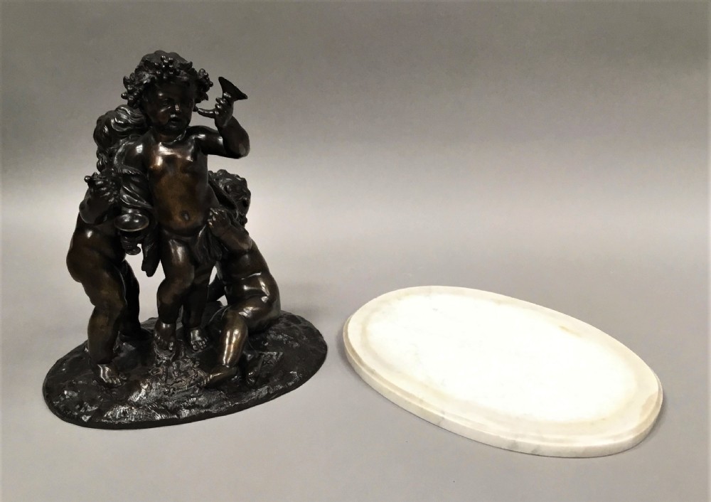 A nineteenth-century dark bronze sculpture depicting three Bacchanalian putti with grape clusters and cups, mounted on a circular white marble plinth. View XV