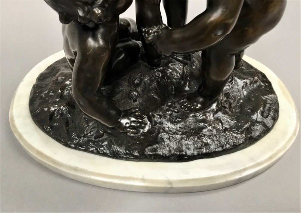 A nineteenth-century dark bronze sculpture depicting three Bacchanalian putti with grape clusters and cups, mounted on a circular white marble plinth. View XVII