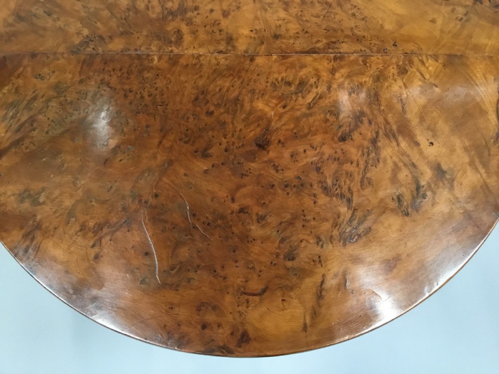 A Regency period tilt-top centre table made from burr yew wood, featuring a circular top displayed vertically on a tripod pedestal base with brass castors. View V