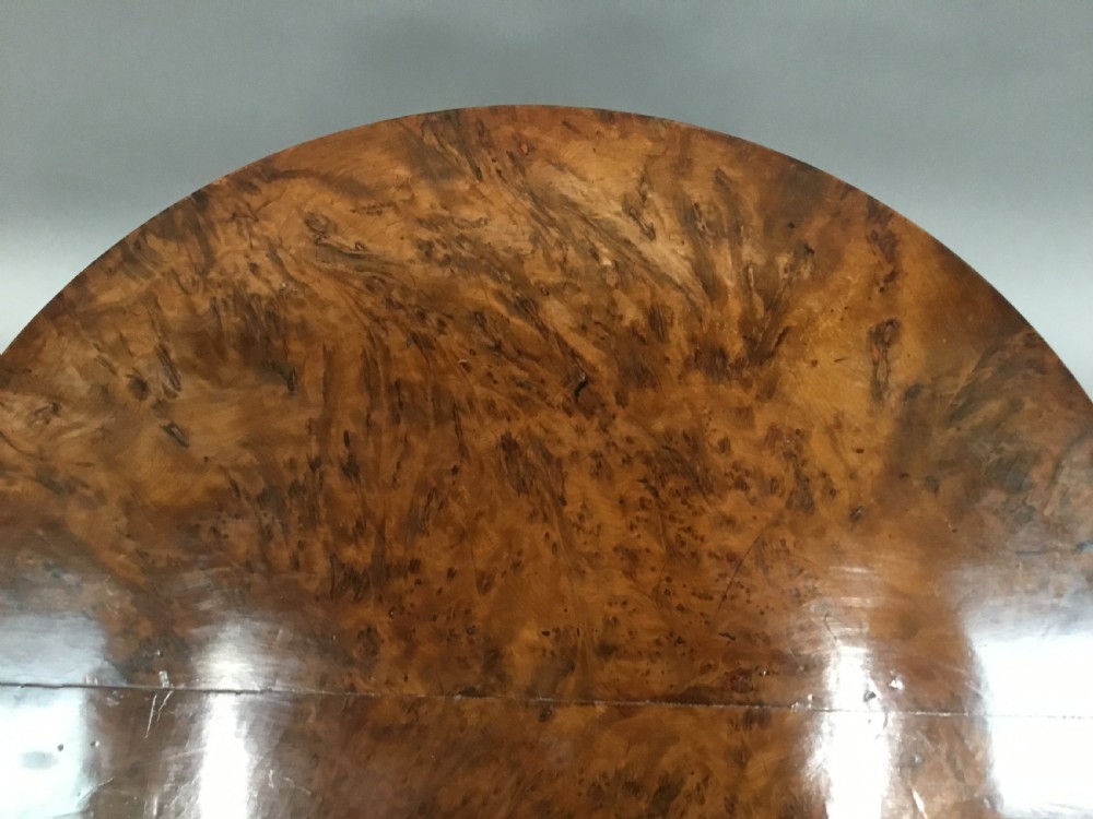 A Regency period tilt-top centre table made from burr yew wood, featuring a circular top displayed vertically on a tripod pedestal base with brass castors. View VI