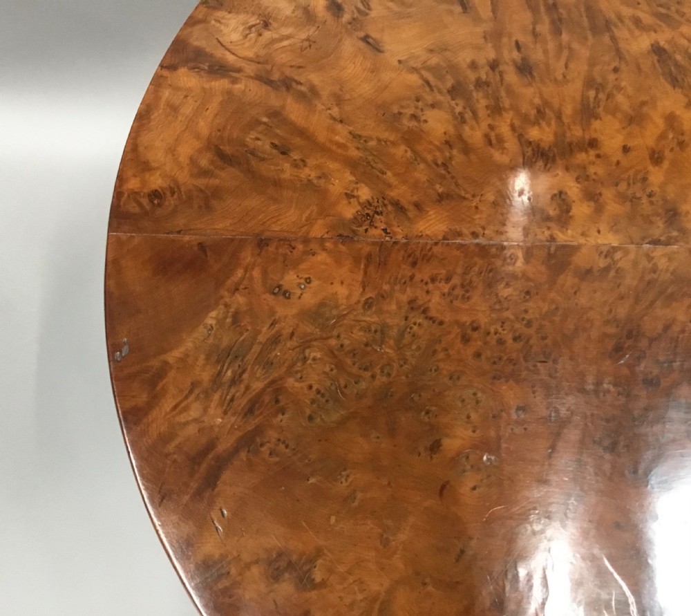 A Regency period tilt-top centre table made from burr yew wood, featuring a circular top displayed vertically on a tripod pedestal base with brass castors. View VII