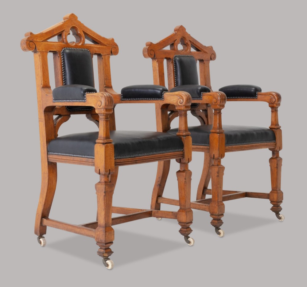 A pair of 19th-century Gothic Revival golden oak open armchairs with black leather upholstery and carved trefoil pediments.