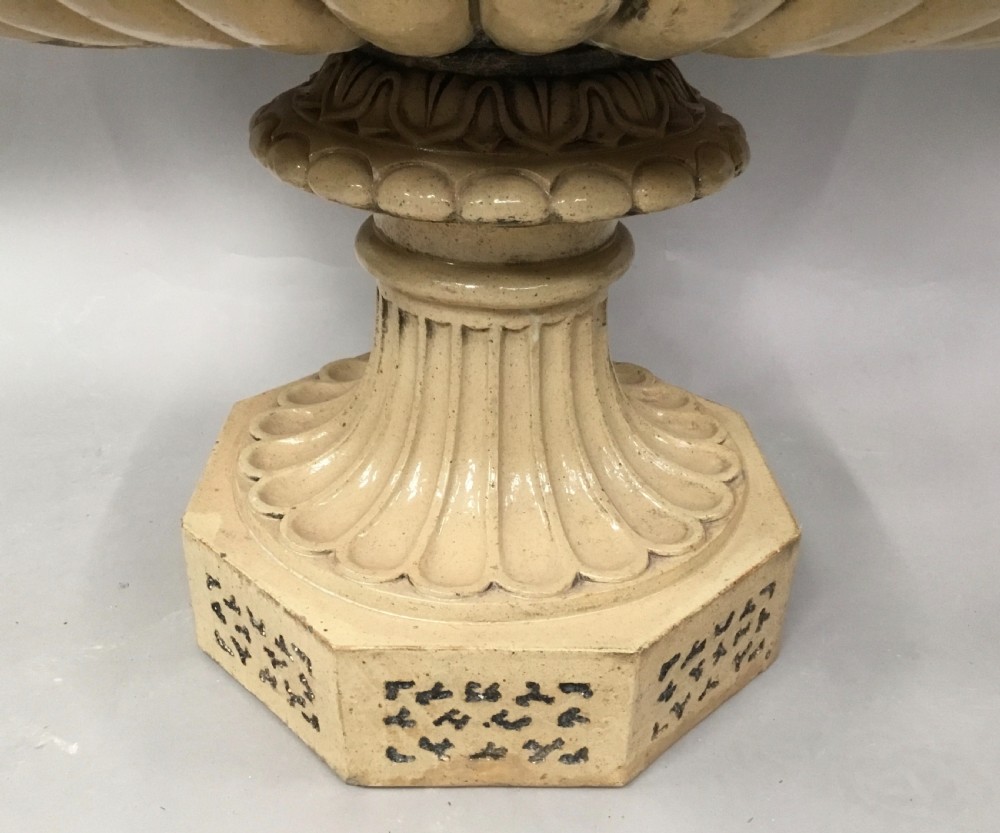 A cream glazed terracotta pedestal urn with a gadrooned lower bowl and an octagonal base featuring incised markings. View IV