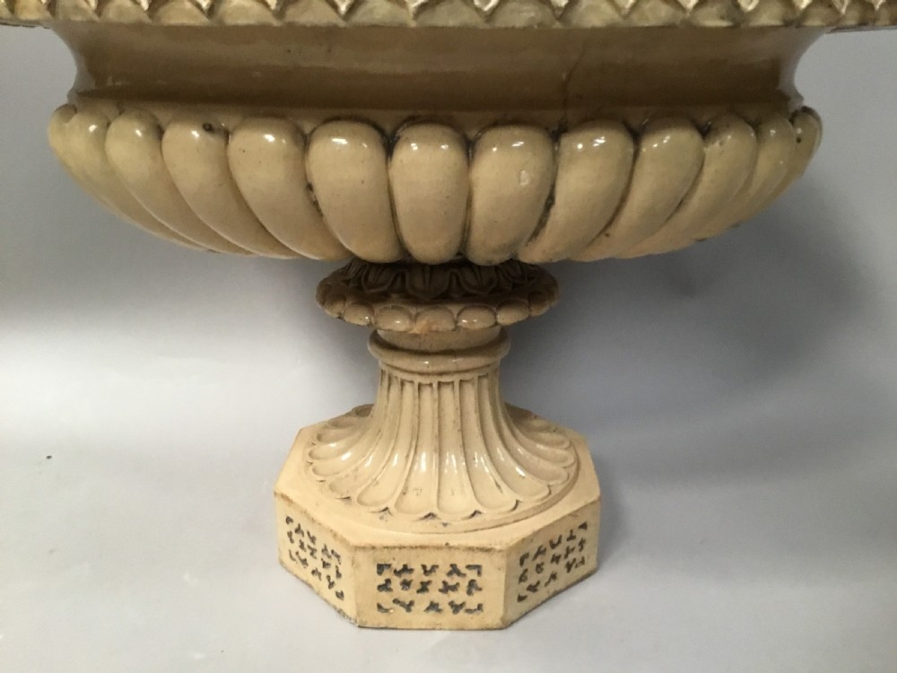 A cream glazed terracotta pedestal urn with a gadrooned lower bowl and an octagonal base featuring incised markings. View XV