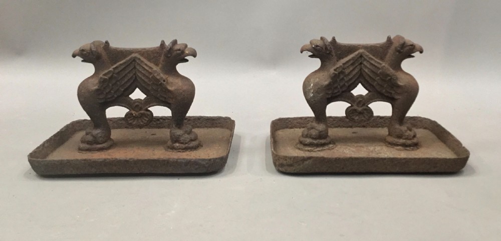 A pair of antique cast iron boot scrapers featuring sculpted griffin side supports and rectangular drip trays with an oxidised surface. View X