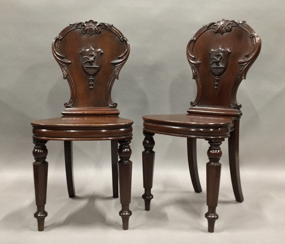 A pair of dark George IV mahogany hall chairs featuring carved backrest scrolls, crests depicting a hippocampus, and turned front legs.