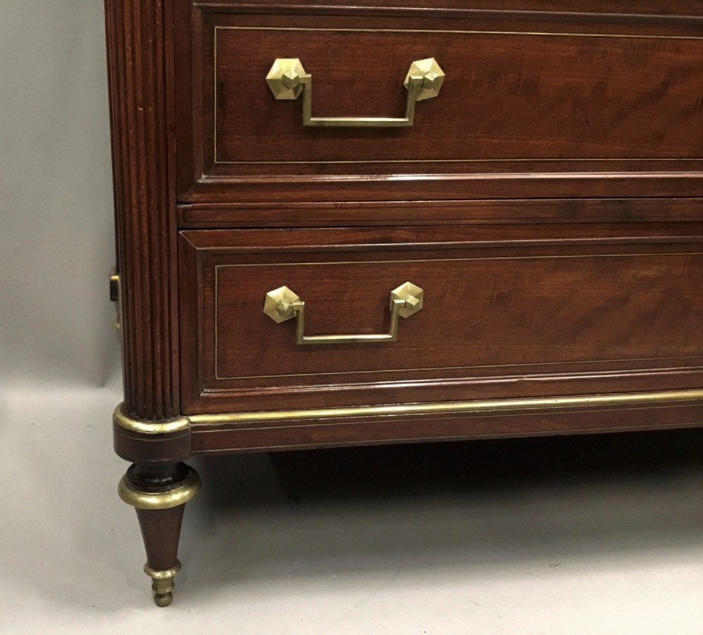 A late 18th-century French Louis XVI neoclassical commode in figured mahogany with a top brass gallery, four drawers, fluted corner stiles, angular brass handles, and turned tapered legs. View IV