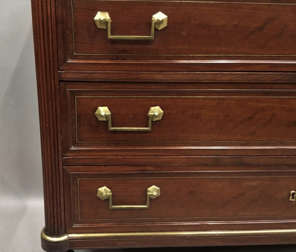 A late 18th-century French Louis XVI neoclassical commode in figured mahogany with a top brass gallery, four drawers, fluted corner stiles, angular brass handles, and turned tapered legs. View XVIII
