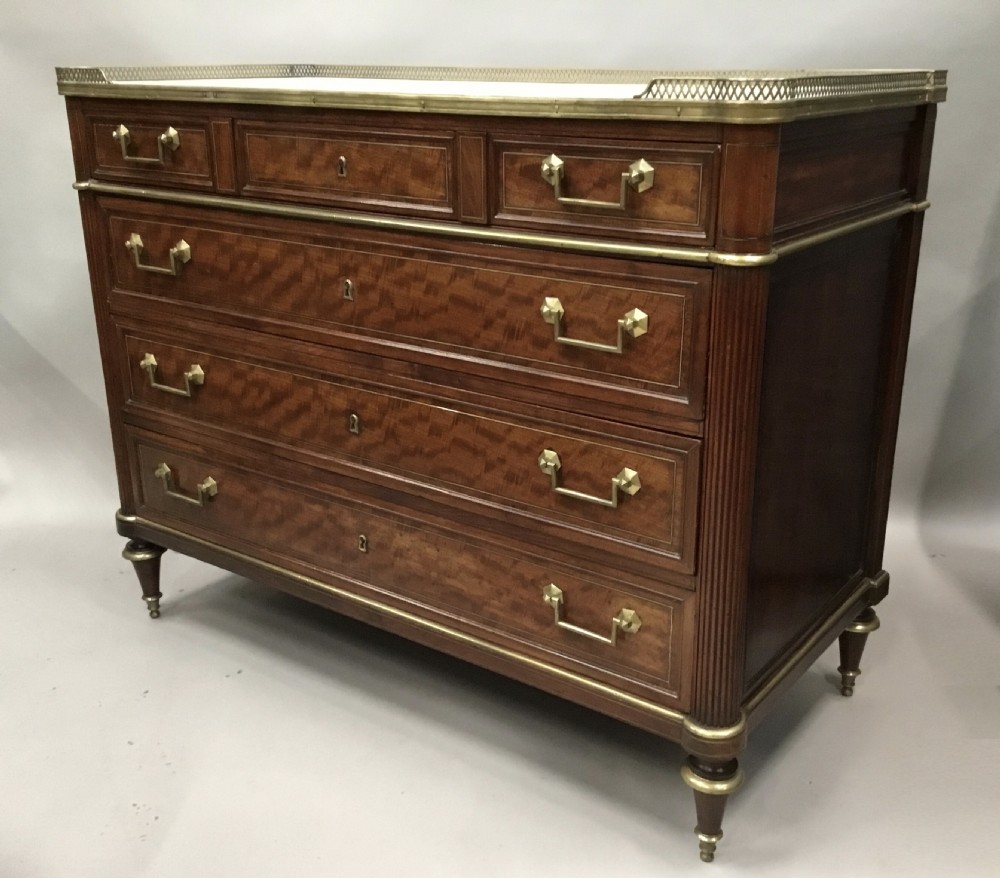A late 18th-century French Louis XVI neoclassical commode in figured mahogany with a top brass gallery, four drawers, fluted corner stiles, angular brass handles, and turned tapered legs.