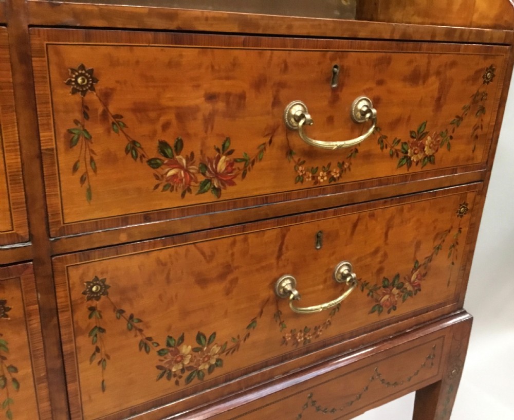 A George III satinwood bookcase cabinet featuring open shelves, painted floral garlands, four drawers with brass drop handles, and square tapered legs. View VIII
