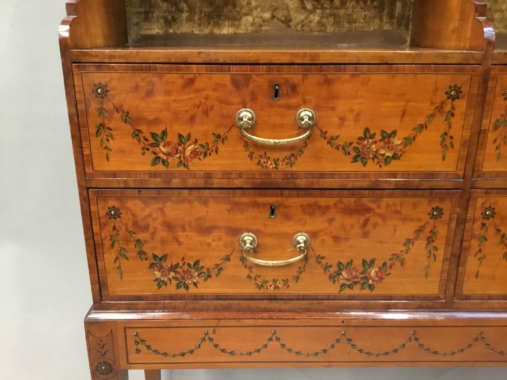 A George III satinwood bookcase cabinet featuring open shelves, painted floral garlands, four drawers with brass drop handles, and square tapered legs. View XX
