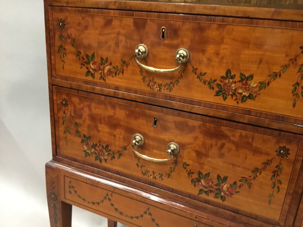 A George III satinwood bookcase cabinet featuring open shelves, painted floral garlands, four drawers with brass drop handles, and square tapered legs. View XXI