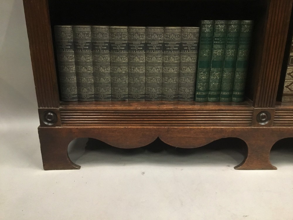 A low Regency mahogany open bookcase featuring two sections with fixed shelving, flanked by fluted pilasters with roundels, set over a shaped apron. View XX