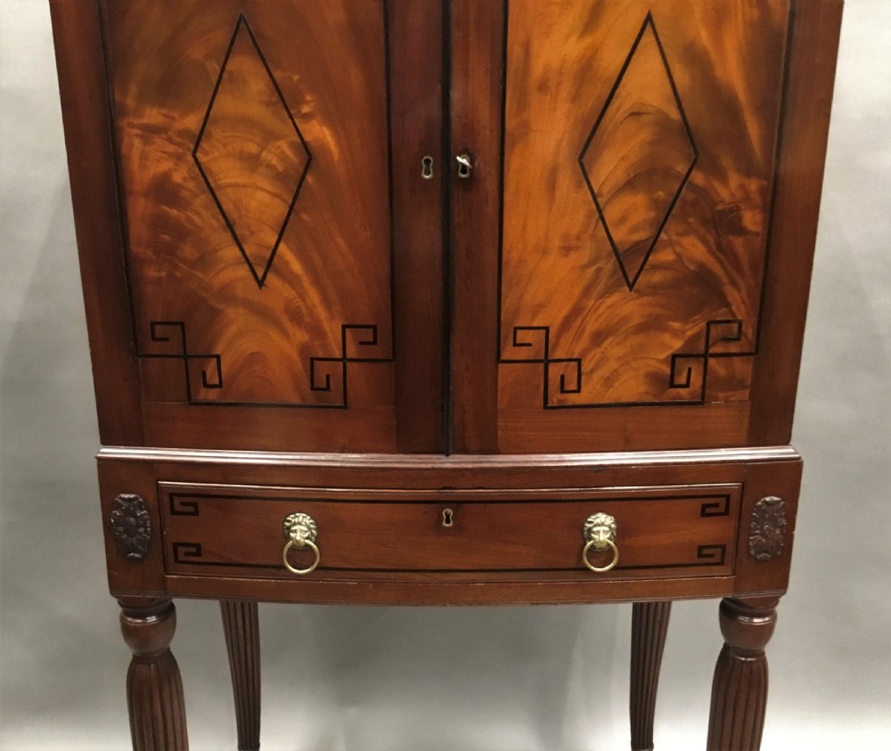A Regency mahogany cabinet on stand featuring flame mahogany door panels with ebony geometric inlay, a single frieze drawer, reeded legs, and brass paw feet. View VI