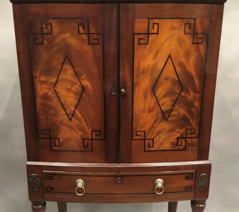 A Regency mahogany cabinet on stand featuring flame mahogany door panels with ebony geometric inlay, a single frieze drawer, reeded legs, and brass paw feet. View XI