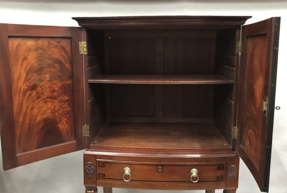 A Regency mahogany cabinet on stand featuring flame mahogany door panels with ebony geometric inlay, a single frieze drawer, reeded legs, and brass paw feet. View XIX