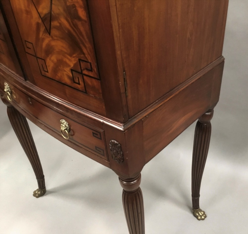 A Regency mahogany cabinet on stand featuring flame mahogany door panels with ebony geometric inlay, a single frieze drawer, reeded legs, and brass paw feet. View XXI