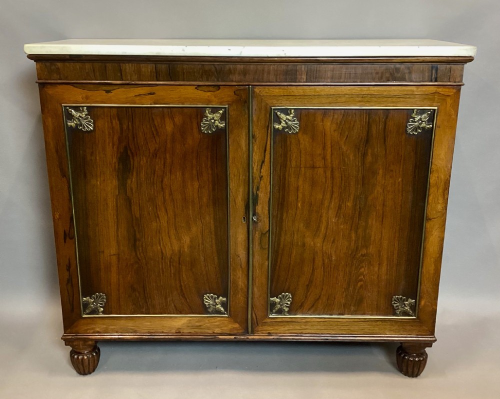A Regency rosewood side cabinet featuring two panelled doors with brass corner mounts, set beneath a white marble top.