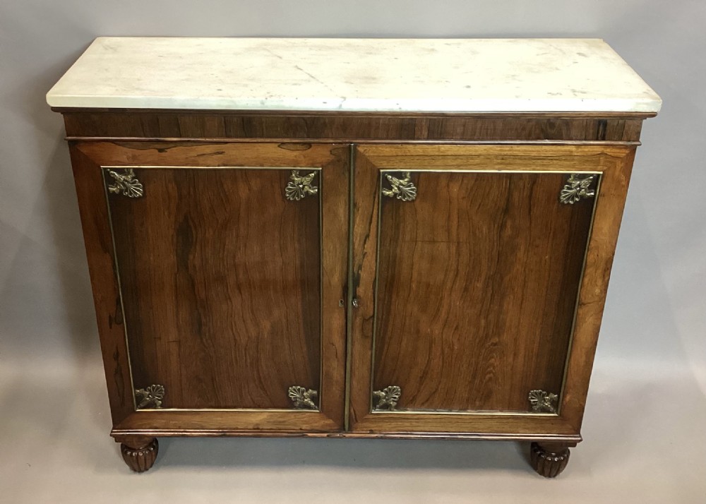 A Regency rosewood side cabinet featuring two panelled doors with brass corner mounts, set beneath a white marble top. View V