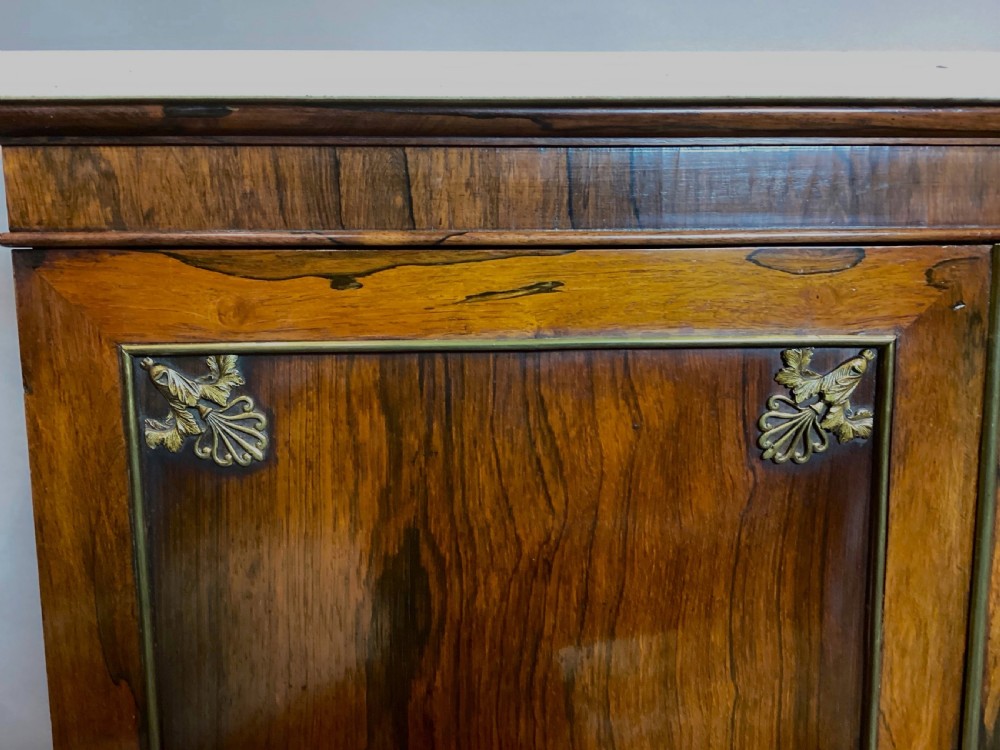 A Regency rosewood side cabinet featuring two panelled doors with brass corner mounts, set beneath a white marble top. View VI