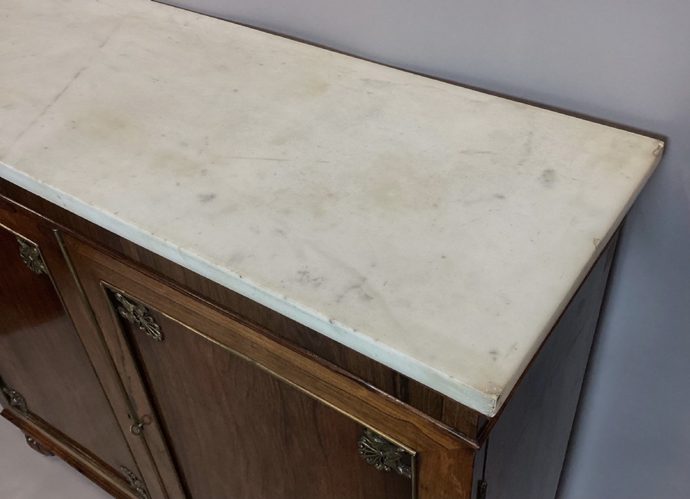 A Regency rosewood side cabinet featuring two panelled doors with brass corner mounts, set beneath a white marble top. View IX