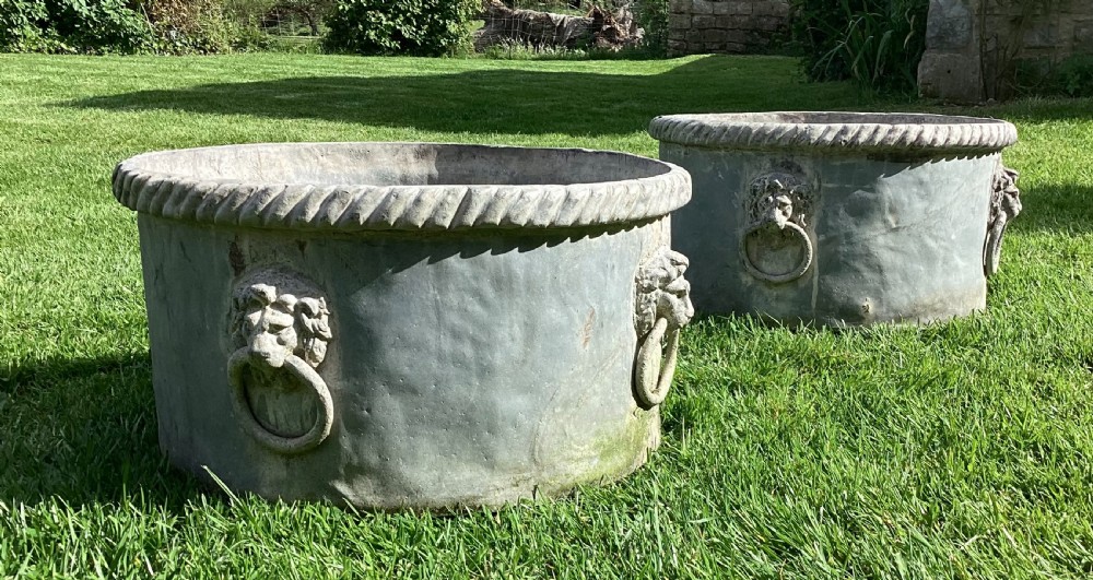A pair of large cylindrical Regency lead garden planters featuring moulded rope-twist rims and relief lion mask ring handles with a weathered grey patina.