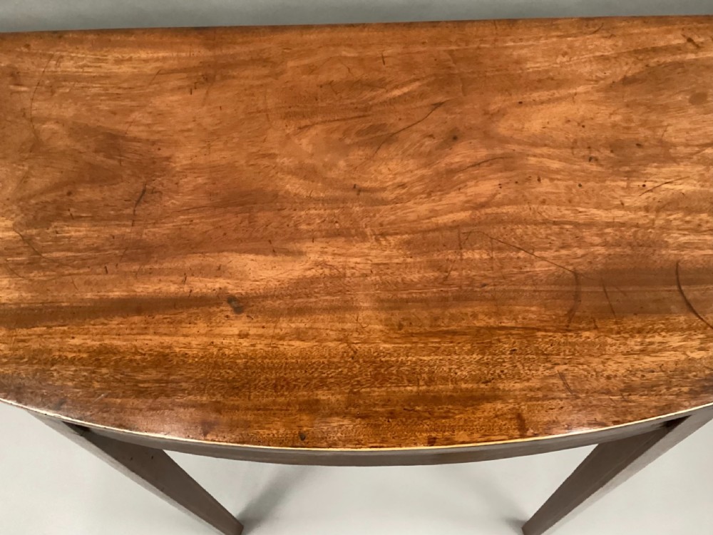 A semi-circular George III style mahogany console table featuring subtle line inlay and four slender square tapered legs. View IX