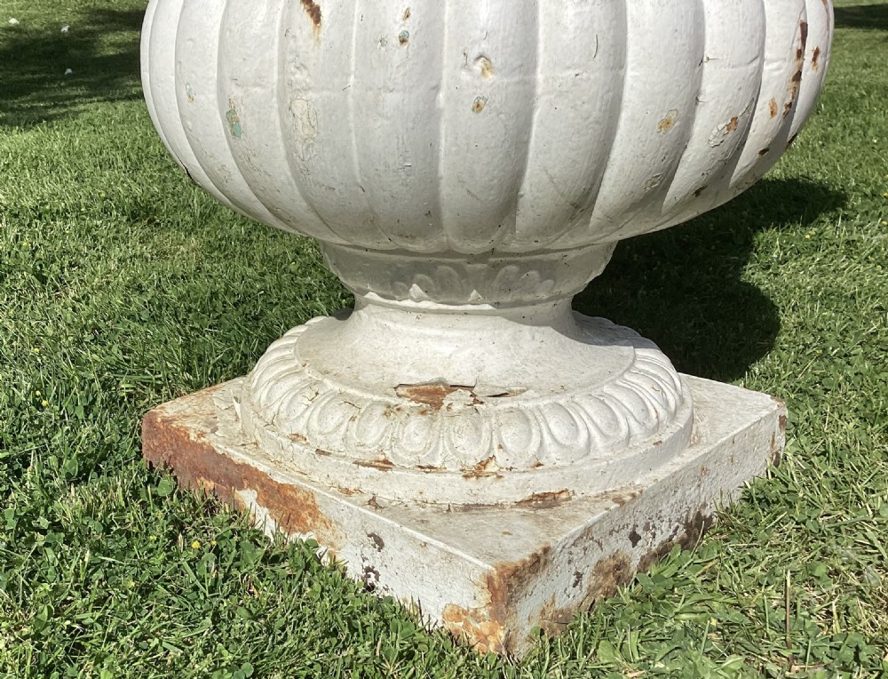 A white painted 19th-century cast iron garden urn with fluted gadrooning, leaf relief carving, side ring handles, and a square pedestal base. View X