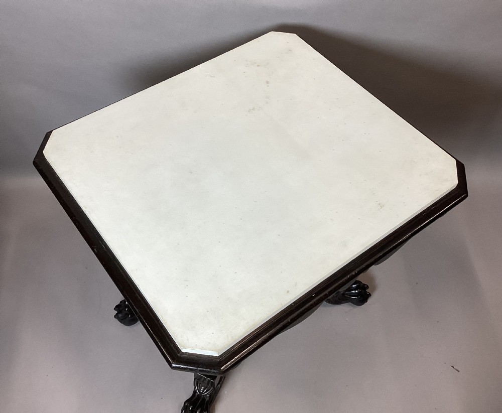 A George IV period ebonised centre table featuring a white rectangular marble top set above a shaped apron, central column pedestal, and quadform base with carved paw feet. View IV