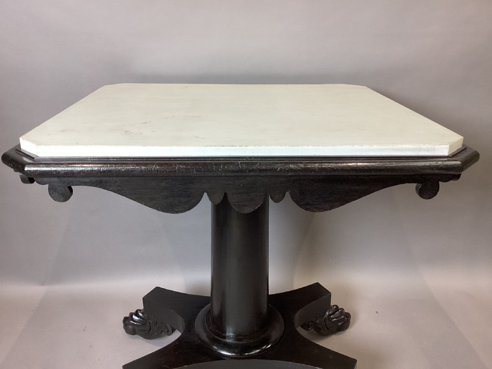 A George IV period ebonised centre table featuring a white rectangular marble top set above a shaped apron, central column pedestal, and quadform base with carved paw feet. View VI
