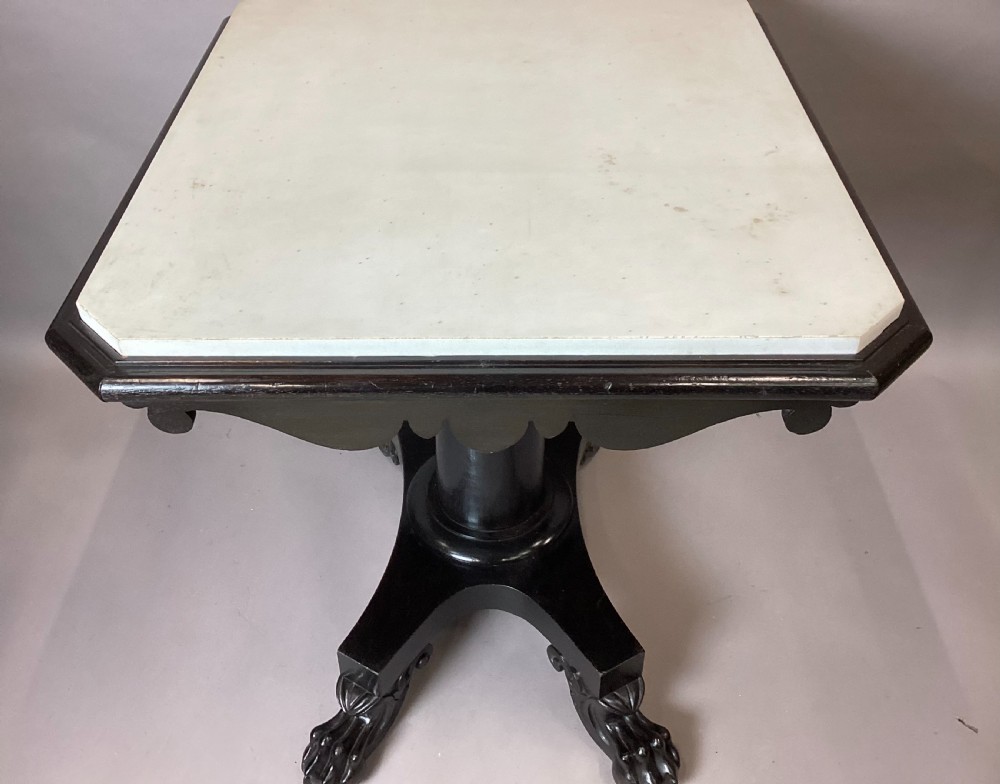 A George IV period ebonised centre table featuring a white rectangular marble top set above a shaped apron, central column pedestal, and quadform base with carved paw feet. View XVIII
