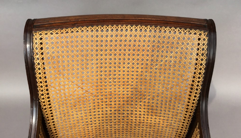 A Regency mahogany bergere library chair with woven cane back, sides, and seat, set on curved legs with brass castors. View XVI
