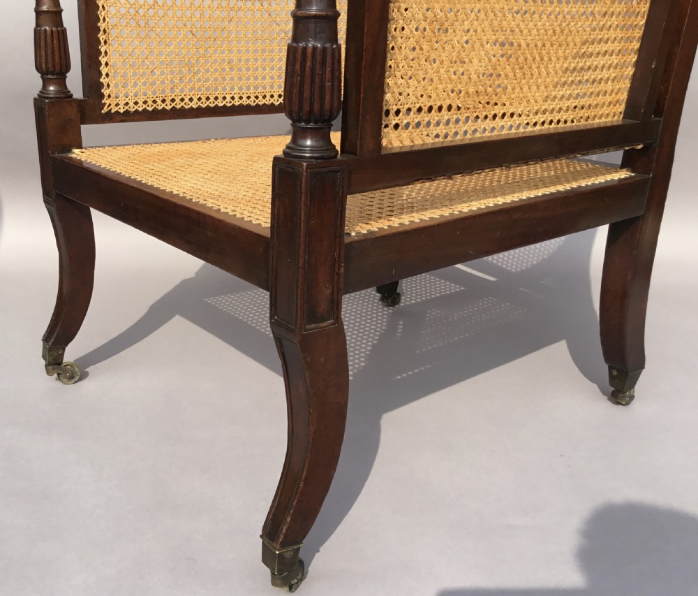A Regency mahogany bergere library chair with woven cane back, sides, and seat, set on curved legs with brass castors. View XVII