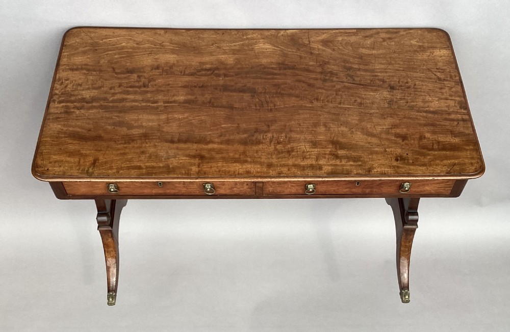 An Irish Regency mahogany centre table with two frieze drawers and brass lion mask handles, resting on end trestle supports with sabre legs and brass castors. View II