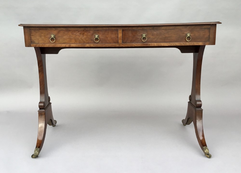 An Irish Regency mahogany centre table with two frieze drawers and brass lion mask handles, resting on end trestle supports with sabre legs and brass castors. View IX