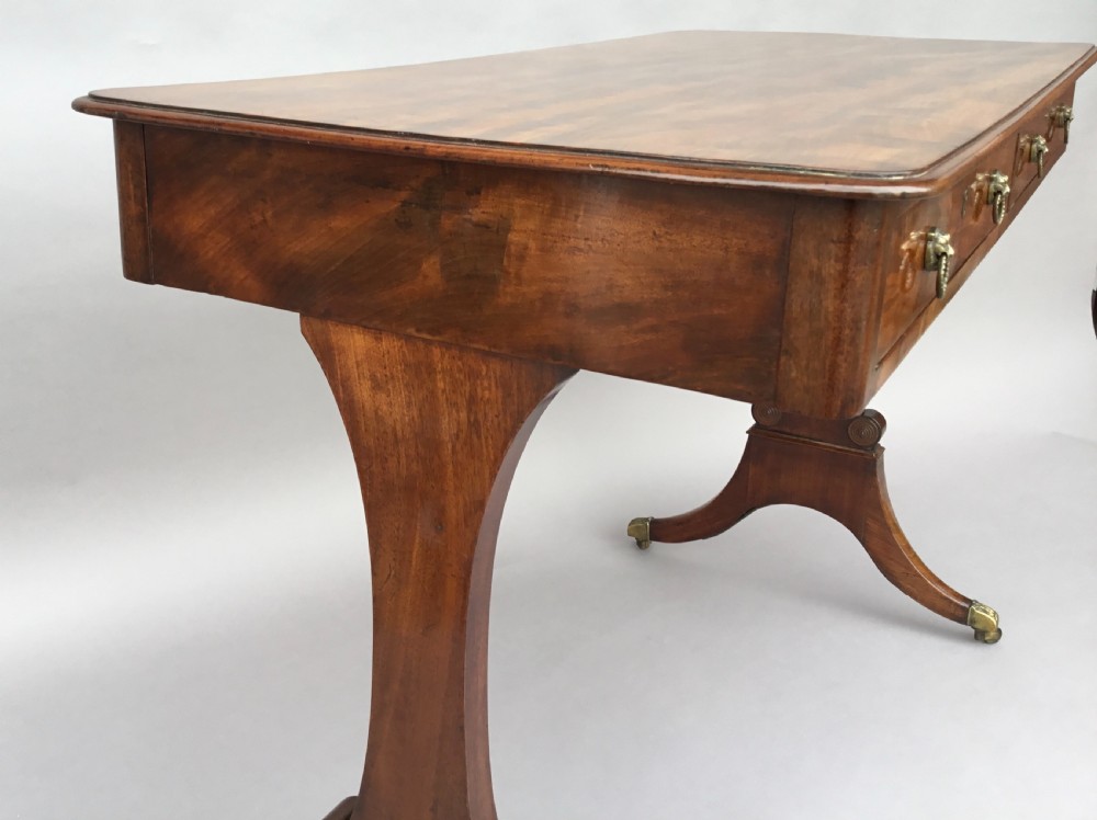 An Irish Regency mahogany centre table with two frieze drawers and brass lion mask handles, resting on end trestle supports with sabre legs and brass castors. View XIV