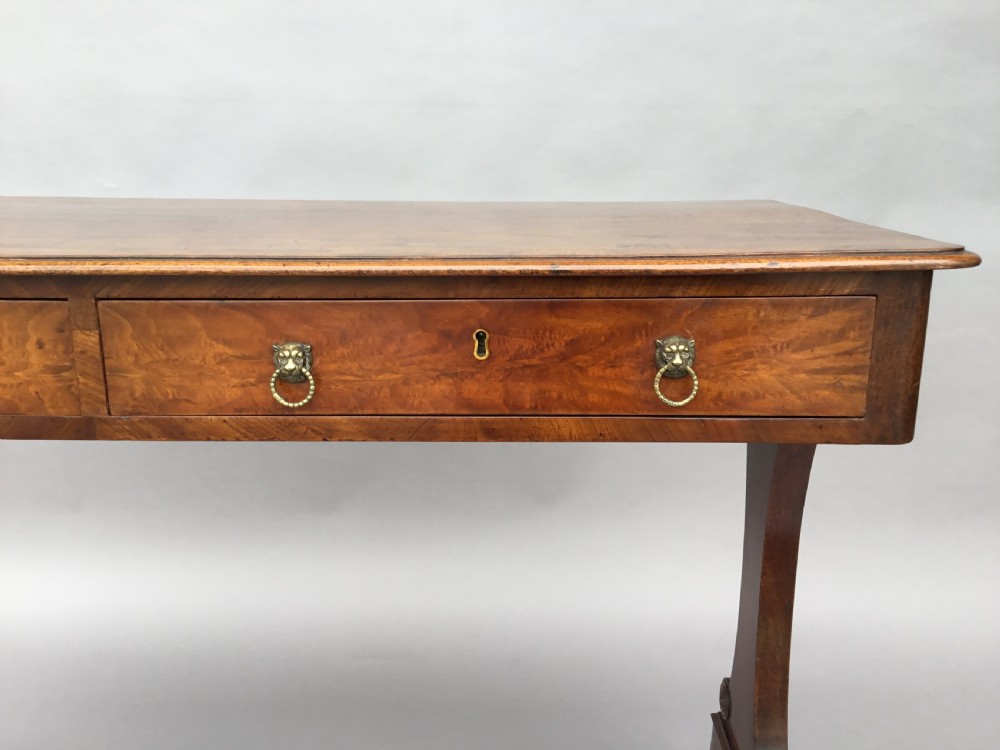 An Irish Regency mahogany centre table with two frieze drawers and brass lion mask handles, resting on end trestle supports with sabre legs and brass castors. View XVI