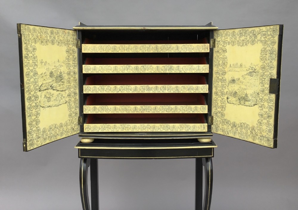 A Regency black lacquered collectors cabinet on stand featuring detailed cream and gilt Chinoiserie penwork illustrations of oriental figures and floral borders. View III