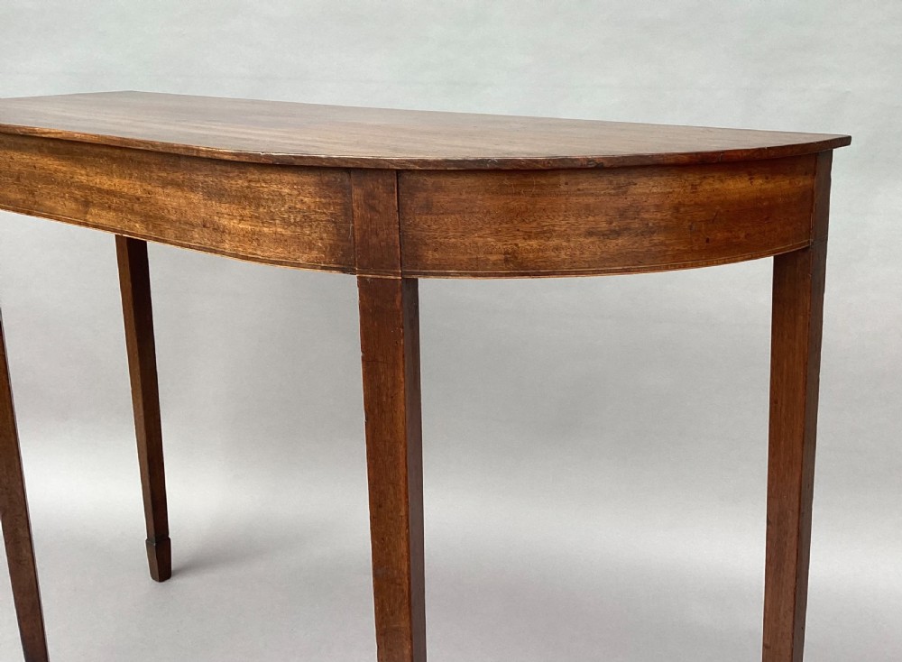 A George III mahogany demilune console table featuring a semi-circular top, plain apron, and four long square tapered legs. View IV