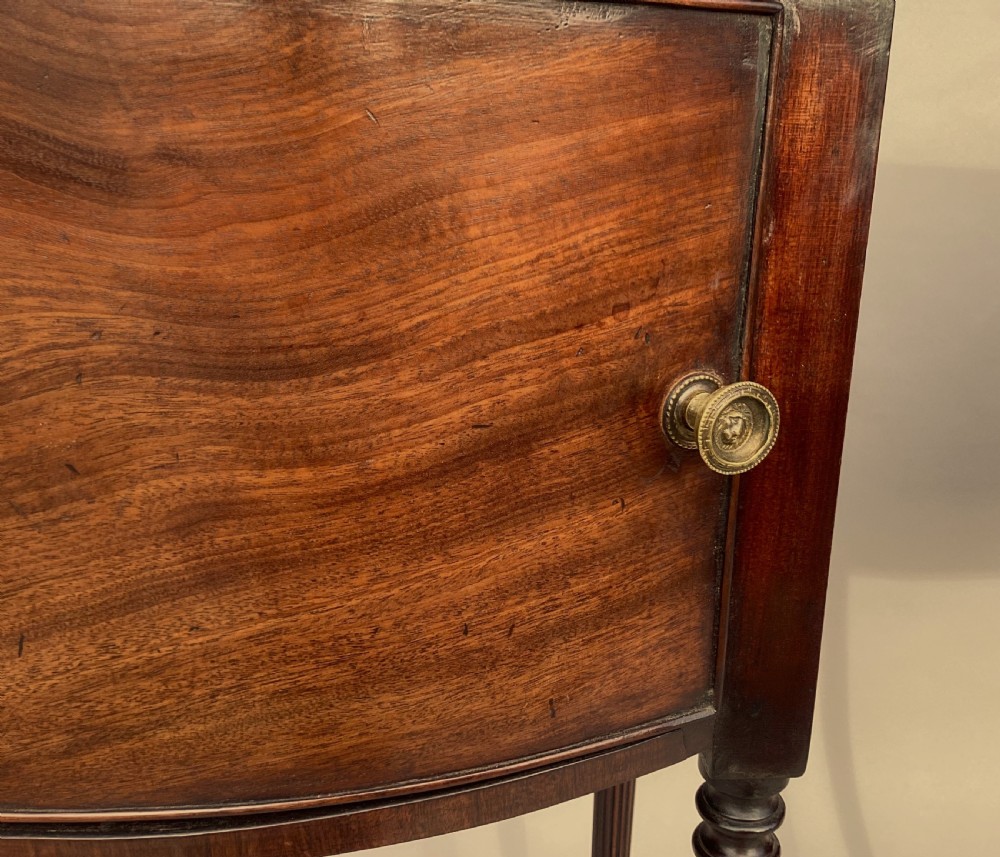 A matching pair of George III mahogany bowfront bedside cabinets standing on slender reeded turned legs with single cupboard doors. View XXI