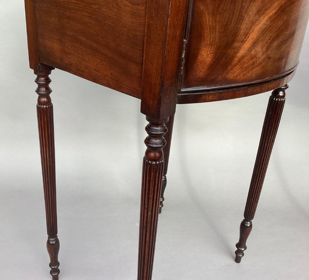 A matching pair of George III mahogany bowfront bedside cabinets standing on slender reeded turned legs with single cupboard doors. View XXIV