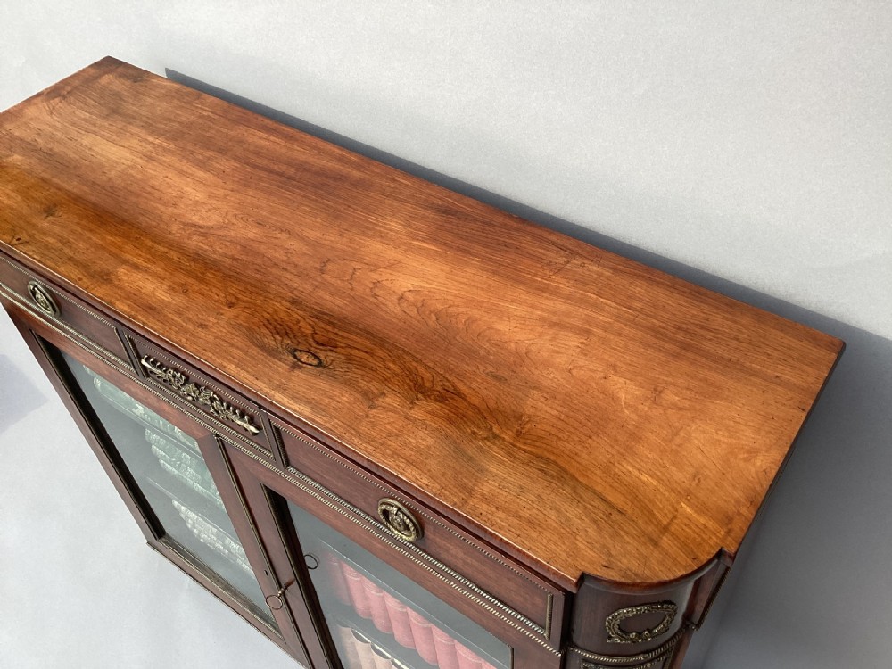 A Regency period dark rosewood side cabinet featuring gilt brass mouldings, decorative wreath appliques, two drawers, glass doors, and brass ball feet. View VI