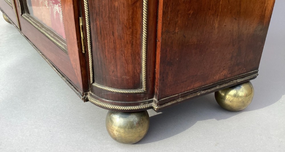 A Regency period dark rosewood side cabinet featuring gilt brass mouldings, decorative wreath appliques, two drawers, glass doors, and brass ball feet. View IX