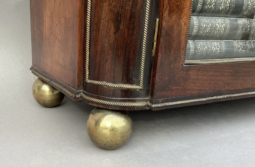 A Regency period dark rosewood side cabinet featuring gilt brass mouldings, decorative wreath appliques, two drawers, glass doors, and brass ball feet. View X