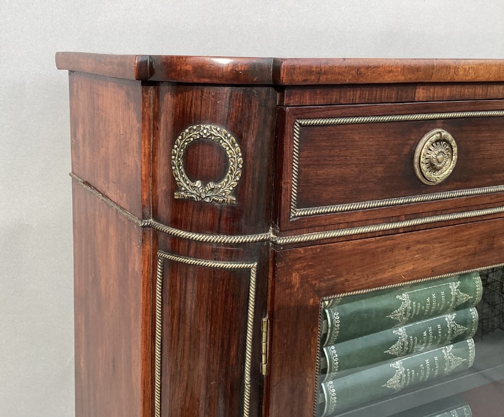 A Regency period dark rosewood side cabinet featuring gilt brass mouldings, decorative wreath appliques, two drawers, glass doors, and brass ball feet. View XI